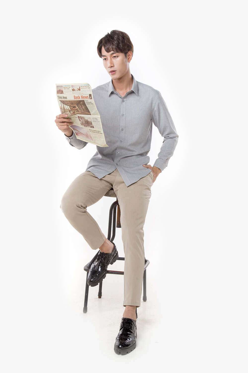 Người mẫu nam ngồi trên ghế, đọc báo trong khi mặc áo sơ mi Café-DriS màu xám và quần kaki màu be